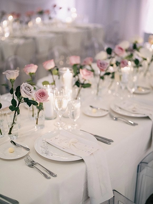 Wedding tablescape with blush rose centerpiece in glass bud vases, pillar candles, menus and place cards on a white tablecloth in soft bokeh light