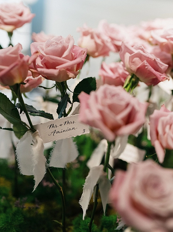 Wedding escort cards tied to pink roses with ribbon and handwritten deckle-edge place cards in soft indoor light on a blurred table setting