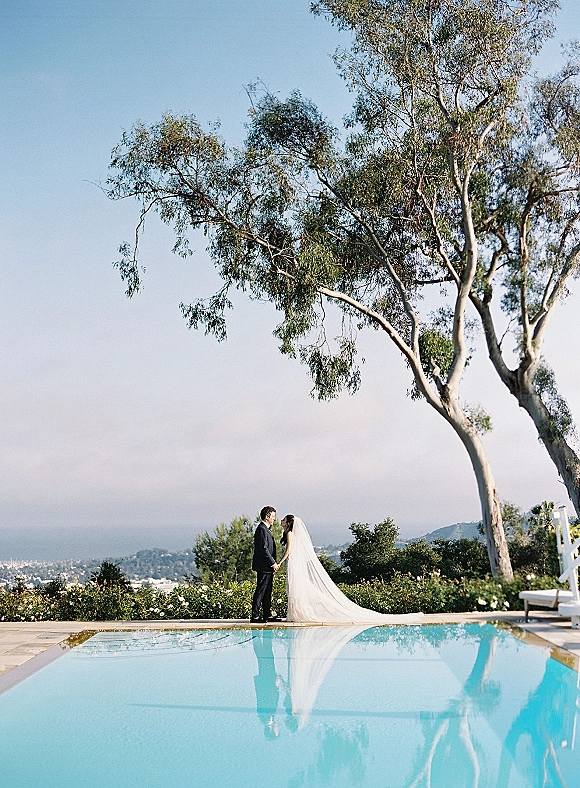 Couple portrait of bride and groom by pool, holding hands with a flowing wedding veil, framed by a large tree and hills under open sky