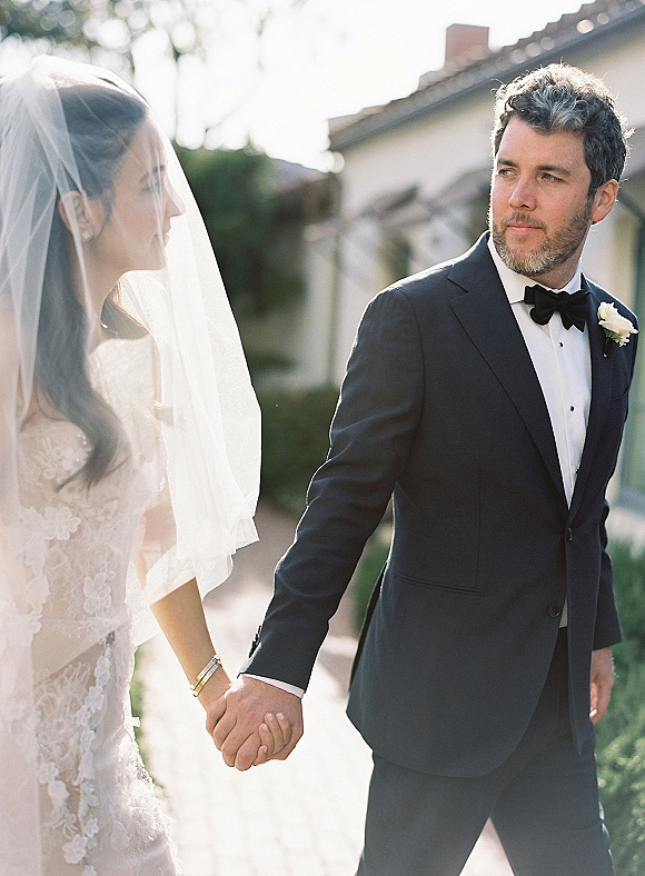 Couple portrait of bride and groom holding hands, her veil flowing beside his black tuxedo, on a sunlit walkway with greenery