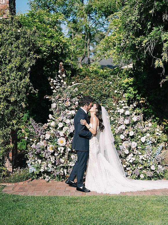 Wedding kiss portrait of the bride and groom kissing under a floral backdrop, her long veil trailing over a brick walkway in a lush garden