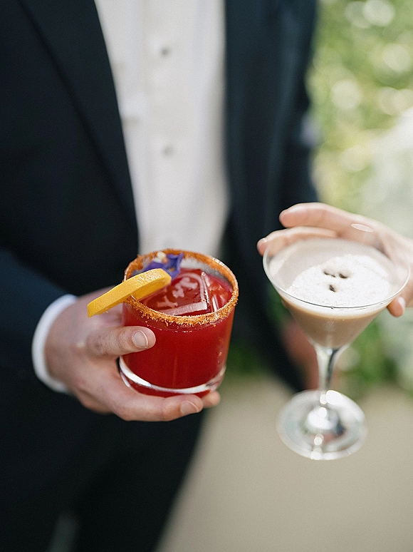 Wedding cocktails held by a tuxedoed groom in two glasses, featuring a red sugar-rim drink and espresso martini with orange garnish, greenery bokeh backdrop