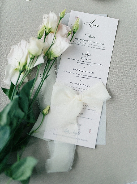 Wedding menu card with wedding menu design in calligraphy, styled beside white flowers and ribbon on a linen tablecloth