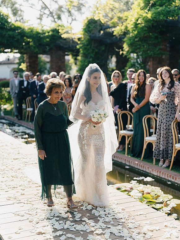 Bride walking down aisle in an outdoor processional, wearing a lace dress and veil, holding bouquet beside a pond with lily pads in sunlight