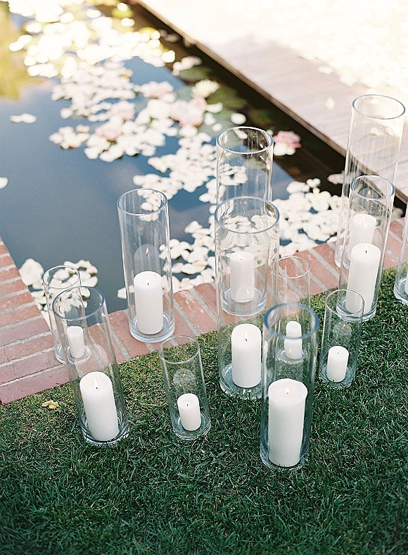 Wedding candle decor with glass hurricane candles and white pillar candles in cylinder vases beside a reflecting pool on a lawn walkway