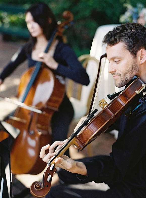 Wedding musicians performing as a wedding string duo with violin and cello, seated in black attire beside a garden path with greenery