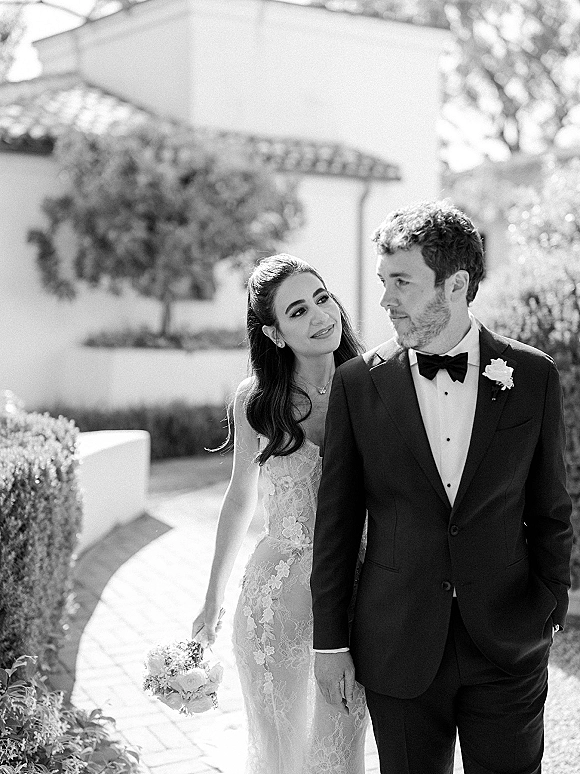 Couple portrait in a garden path, bride in strapless lace dress holding bouquet behind tuxedoed groom under sunlight and hedges