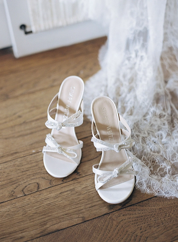 Bridal shoes in white wedding heels with rhinestone bow straps beside a lace wedding dress on a wood floor near a white door