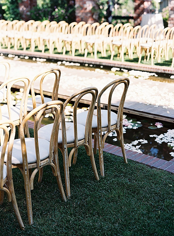 Ceremony seating with outdoor ceremony chairs in neat rows facing a raised aisle platform over a pond with lily pads and petals