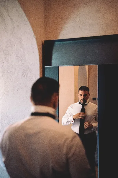Groom getting ready, tying black necktie over a white dress shirt, seen in a mirror reflection beside an arched doorway indoors