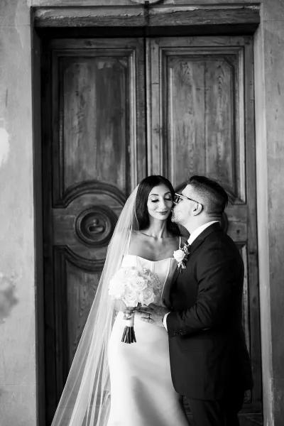 Wedding couple portrait of groom kissing the bride’s cheek as she holds a rose bouquet, standing by rustic wooden double doors in stone doorway