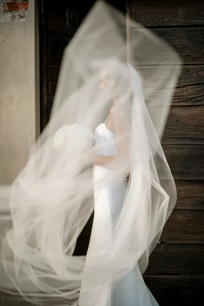 Bridal portrait of a bride with veil blowing over her face, holding a white rose bouquet in a doorway against a wood wall backdrop