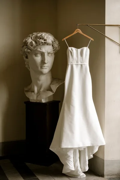 Wedding dress in simple satin hangs on a wooden hanger beside a sculpture bust on a pedestal near a beige stair railing