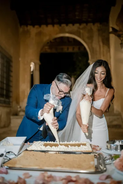 Wedding cake cutting as bride and groom decorate a white sheet cake with piping bags under a stone arch in an outdoor courtyard at night