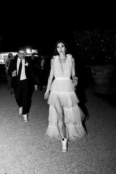 Reception exit moment as bride in tulle dress and sneakers walks with tuxedoed groom holding a drink along a lit gravel path at night