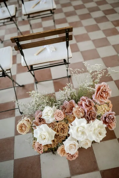 Wedding aisle flowers in blush and white rose arrangements with baby's breath and greenery beside wooden chairs on a checkered tile floor