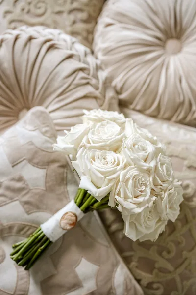 Bridal bouquet of white roses with a ribbon wrap resting on a patterned neutral sofa, styled as an elegant wedding detail close-up
