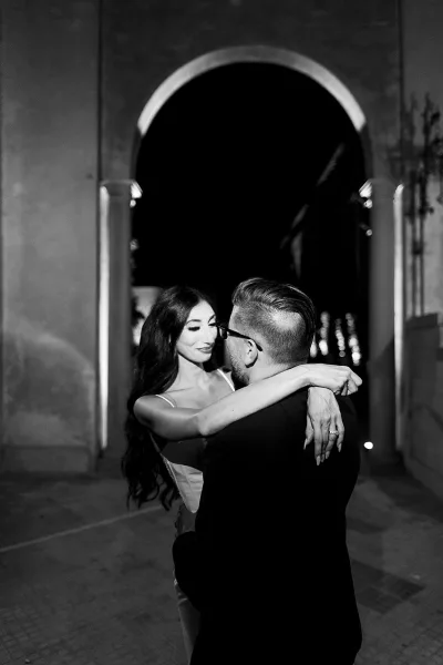 Wedding couple portrait in black and white as bride hugs groom in glasses under an arched doorway along a courtyard walkway at night