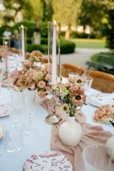 Reception tablescape with an outdoor reception table set with blush runner, taper candles, floral centerpieces, wicker chairs in garden greenery