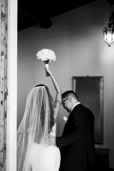 Wedding couple portrait in a black and white wedding photo, bride lifting a white rose bouquet overhead as they walk through a doorway indoors