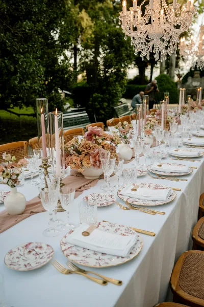 Reception tablescape with outdoor wedding reception table details, blush runner, rose centerpieces, taper candles, and crystal chandelier amid greenery