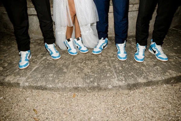 Wedding sneakers lined up in matching blue and white pairs with dress pants and wedding dress hem on stone steps beside a stone wall