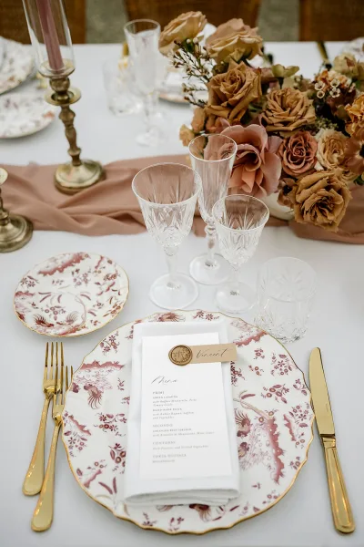 Reception tablescape with patterned china and gold flatware, crystal stemware, wax-seal place cards, and brass taper candles on white linens