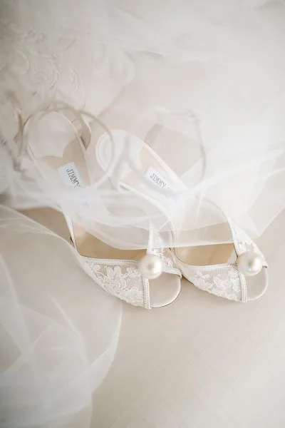 Bridal shoes with pearl wedding heels and lace details, ankle straps and tulle fabric arranged on a light wood floor flatlay