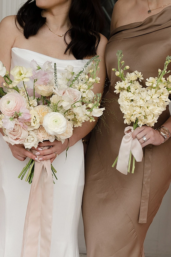 Bridal bouquet and pastel wedding bouquet held by bride in white dress beside bridesmaid in one-shoulder satin dress, blush ribbons trailing indoors