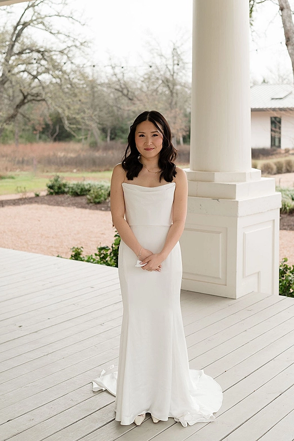 Bridal portrait of a bride in a strapless wedding dress with chapel train, hands clasped on a white porch with columns and trees behind