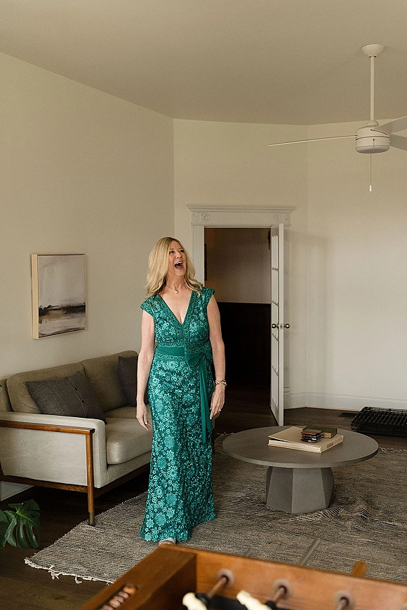 Wedding guest portrait of a woman in a green lace maxi dress with waist sash, standing in a bright living room by a sofa and wall art