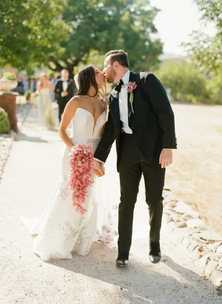 Wedding kiss portrait of bride and groom kissing while holding hands on an outdoor tree-lined path, her veil and pink bouquet flowing