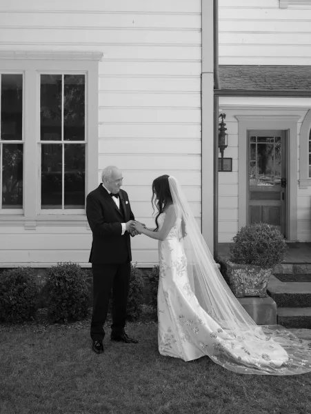 First look moment as father sees bride in embroidered gown and long veil, holding hands on white house porch steps with shrubs