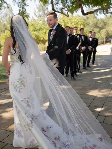 First look moment as bride taps groom’s shoulder, veil trailing behind, groomsmen lined up on a tree-shaded garden walkway