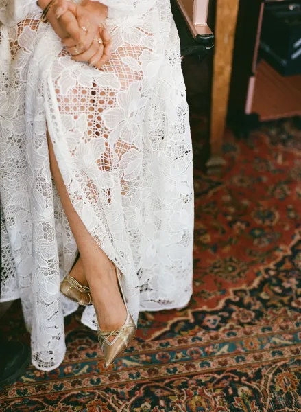 Wedding dress detail with lace wedding dress texture, bridal rings and gold ankle-strap heels arranged on a vintage rug by a chair