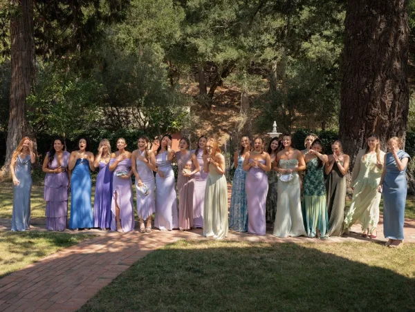 Bridesmaid group photo with bridesmaids reacting in pastel dresses, holding handheld fans and heels on a garden lawn by a brick walkway