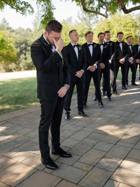 Groom portrait capturing an emotional groom moment as he wipes tears in a black tuxedo and boutonniere beside groomsmen on a tree-lined lawn