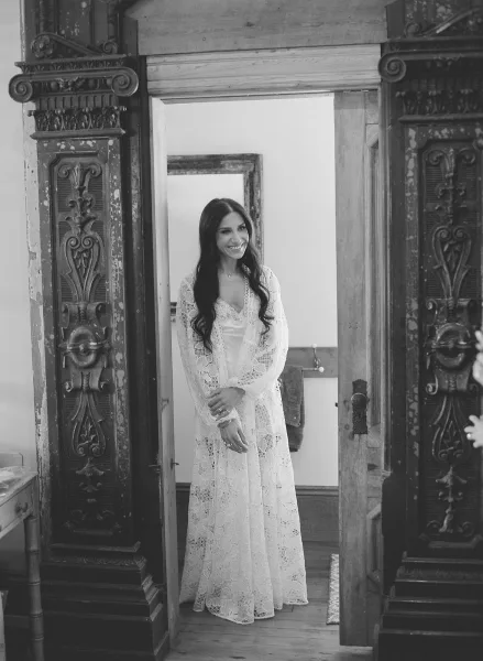 Bridal portrait in black and white of a bride smiling in a doorway, wearing a long sleeve lace gown with a necklace by ornate wood doors