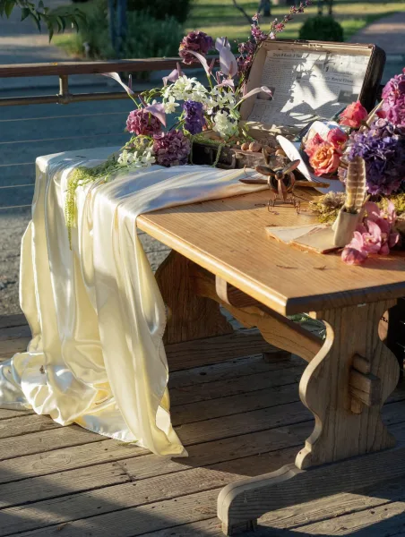 Wedding guestbook table with a vintage suitcase and pen beside purple hydrangea florals on a draped wooden deck welcome display outdoors