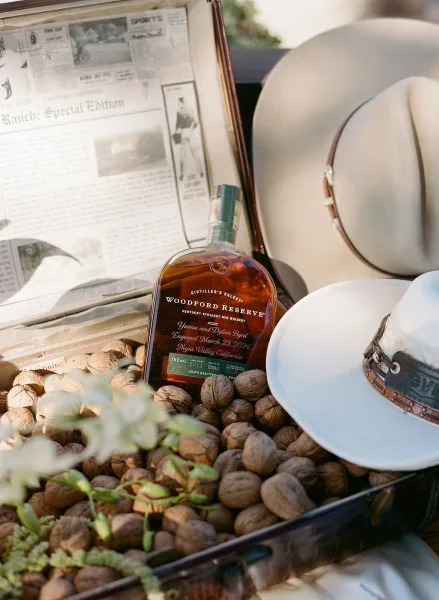 Groom accessories flat lay with whiskey bottle and cowboy hat, styled with a newspaper, suitcase, walnuts, and greenery in soft sunlight