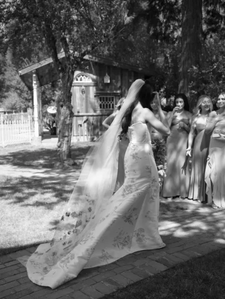 Bride entrance with a long veil and floral appliqué gown as she walks down a brick path, bridesmaids lined up beside garden trees