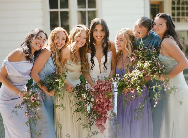 Bridesmaid group photo with bride and bridesmaids in mismatched dresses holding pink bouquets and greenery outside a white house in daylight