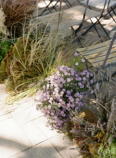 Ceremony aisle florals with purple wildflower clusters, moss and ornamental grasses lining a stone paver walkway beside wooden benches