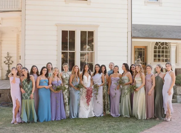 Bridesmaid group photo with bride in gown and mixed bridesmaid dresses holding garden-style bouquets by a white house and brick walkway