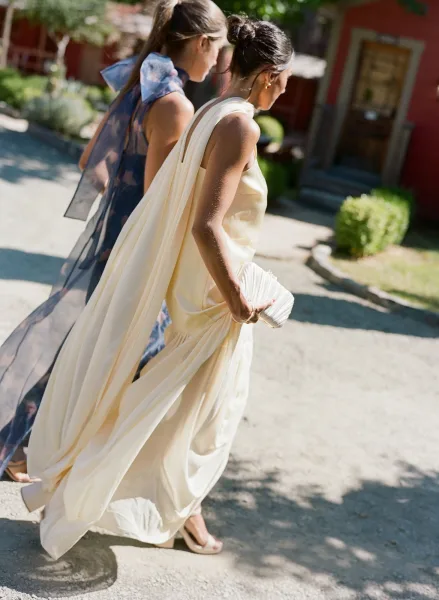 Bride walking with bridesmaid walking along a garden walkway, long veil flowing behind her as she holds a white pleated clutch