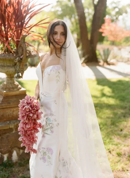 Bridal portrait of a bride holding a cascading pink bouquet in a strapless floral embroidered gown, wedding veil in a sunlit garden