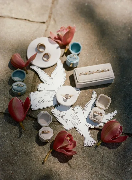 Wedding rings in a wedding ring flat lay with diamond ring and gold bands on a jewelry dish beside pink tulips on sunlit stone pavement