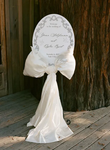 Wedding welcome sign with calligraphy lettering on an oval board, a white fabric bow, set on a metal stand by rustic wood siding