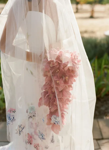 Bridal bouquet with pink orchid bouquet cascading under a sheer veil, tied with ribbon over a floral wedding dress on a stone path
