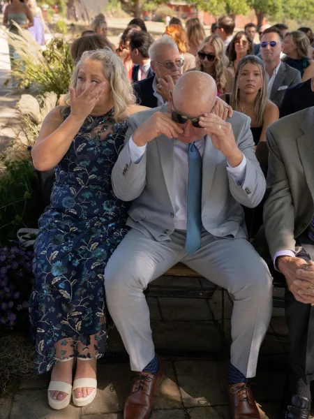 Wedding guest reaction as an older couple in sunglasses and a navy floral dress sit on a wooden bench, teary-eyed in sunlit trees